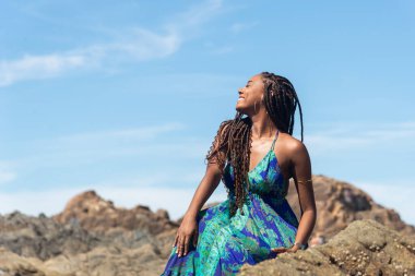 Güzel bir kadın, saç örgüleri ve mavi elbiseleriyle kumsalda güneşlenen kayalarda oturuyor ve mavi gökyüzüne karşı poz veriyor..