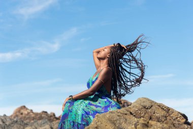 Mavi elbiseli güzel bir kadın kumsalda kayalıklarda güneşleniyor ve mavi gökyüzüne karşı örgülerine dokunuyor..