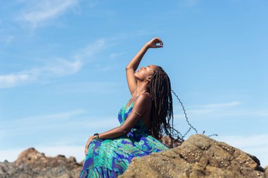 Mavi elbiseli güzel bir kadın kumsalda kayalıklarda güneşleniyor ve mavi gökyüzüne karşı örgülerine dokunuyor..