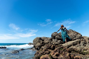 Mavi elbiseli ve örgülü saçlı güzel bir kadın kumsalın tepesinde oturmuş mavi gökyüzüne karşı güneşleniyor..