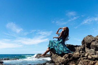 Mavi elbiseli ve örgülü saçlı güzel bir kadın kumsalın kenarında oturmuş mavi gökyüzüne karşı güneşleniyor..
