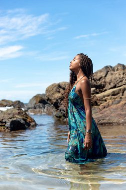 Mavi elbiseli ve örgülü saçlı güzel bir kadın sahilde suyun üzerinde duruyor mavi gökyüzüne karşı poz veriyor..