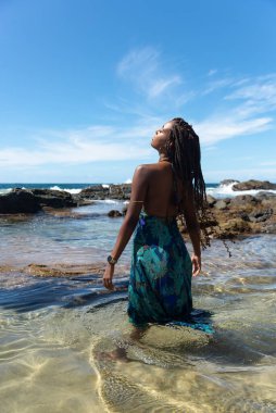 Mavi elbiseli ve örgülü saçlı güzel bir kadın sahilde suyun üzerinde duruyor mavi gökyüzüne karşı poz veriyor..
