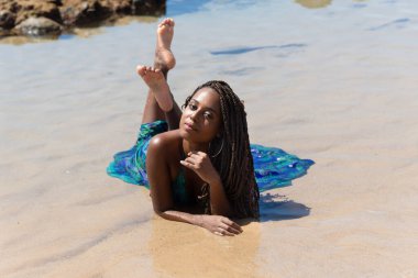 Saçları örgülü ve mavi elbiseli güzel bir kadın plajın kenarında yatıyor. Arka planda kayalara karşı fotoğraf için poz veriyor..