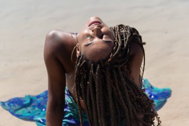 Mavi elbiseli, saçları örgülü, sırtı kapalı bir şekilde kumsalda oturan güzel bir kadın. Seyahat eden mutlu kişi.