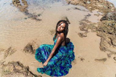 Saçları örgülü güzel bir kadın kumsalda uzun mavi bir elbiseyle güneşleniyor. Seyahat eden mutlu kişi.