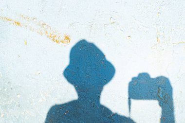 Duvarda kimliği belirsiz bir kişinin gölgesi. Şapka takıyor ve elinde profesyonel bir fotoğraf makinesi tutuyor. Doku.