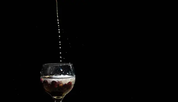Drops of milk falling from above into glass wine glass with milk and grapes inside. Isolated on black studio background.