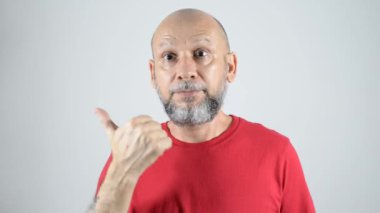 Kırmızı tişörtlü kel sakallı adam sağ tarafa baş parmak işareti yapıyor. Gri arkaplanda izole.
