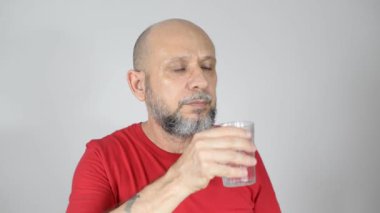 Kırmızı tişörtlü, kel ve sakallı sakin ve sakin bir adam Amerikan bardağından su içiyor. Gri arkaplanda izole.