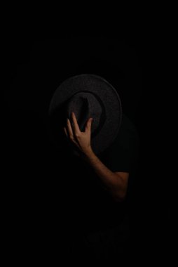 Suratının önünde şapka tutan gizemli bir adam kameraya poz veriyor. Stüdyo portresi. Yapay dumanlı koyu arkaplan.