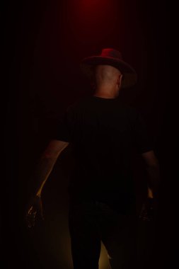 Sırtı kameraya dönük duran şapkalı gizemli adam. Stüdyo portresi. Yapay dumanlı kırmızı koyu arkaplan.