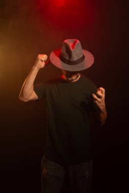 Fotoğraf için poz veren şapkalı gizemli adam. Stüdyo portresi. Yapay dumanlı kırmızı koyu arkaplan.