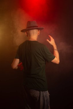 Mysterious man wearing hat standing with his back to the camera. Studio portrait. Red dark background with artificial smoke. clipart