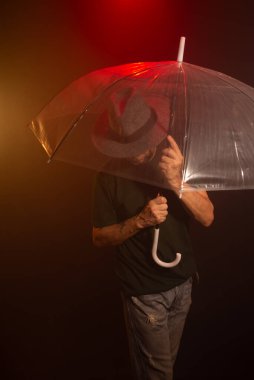 Mysterious man wearing hat standing with a transparent umbrella. Studio portrait. Red dark background with artificial smoke. clipart