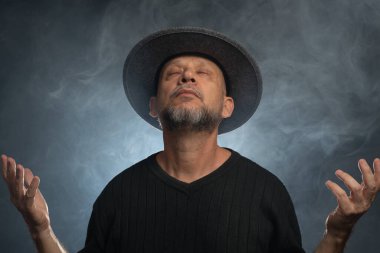 Sakallı, şapkalı ve kameraya poz veren orta yaşlı bir adam. Dumanla dolu arka planda izole edilmiş.