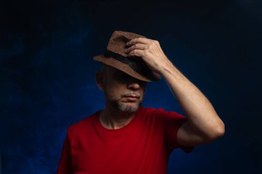 Kırmızı gömlekli, sakallı ve gözlüklü kahverengi şapkalı bir adam. Stüdyoda fotoğraf. Yapay dumanlı mavi stüdyo arka planı.
