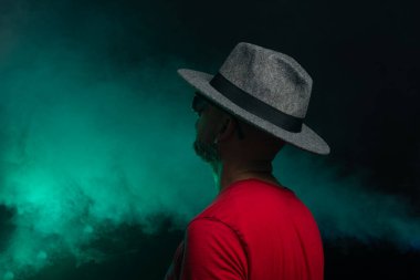 Sakallı adam şapka takıyor ve fotoğraf için poz veriyor. Stüdyoda portre. Yapay dumanlı yeşil arkaplan.