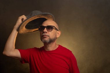 Gözlüklü kel sakallı adam kafasına şapka takıyor. Stüdyo portresi. Yapay dumanla renkli arkaplan.