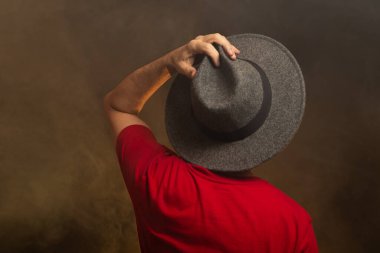 Sırtı kameraya dönük, şapkalı ve fotoğraf için poz veren orta yaşlı sakallı bir adam. Yapay dumanlı renkli stüdyo arkaplanı.