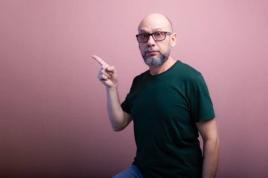 Reçeteli gözlüklü sakallı kel adam parmağını yana doğru gösteriyor. Somon renkli arka planda izole edilmiş.