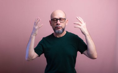 Kel, sakallı, gözlüklü, iki eli başının yanında, gözleri kapalı bir adam portresi. Somon renkli arka planda izole edilmiş.