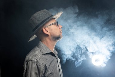 Sakallı, şapkalı ve kameraya poz veren orta yaşlı bir adam. Dumanla dolu arka planda izole edilmiş.
