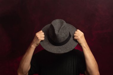 Gizemli, orta yaşlı bir adam iki eliyle şapkasını kafasına dayamış. Stüdyoda fotoğraf. Kırmızı gül renginde yapay duman arka planı.