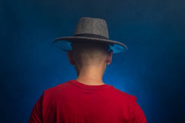 Kırmızı gömlekli, sakallı, gözlüklü, şapkalı, sırtı kameraya dönük bir adam. Stüdyoda fotoğraf. Yapay dumanlı mavi stüdyo arka planı.