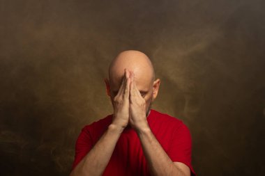 Kel sakallı, yüzünde elleri olan kırmızı tişörtlü bir adam. Stüdyo portresi. Yapay dumanlı sarı arkaplan.