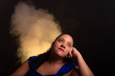 Mavi bluz giyen, fotoğraf için poz veren güzel bir kadın sol tarafa bakıyor. Siyah arkaplanda dumanla izole edilmiş.