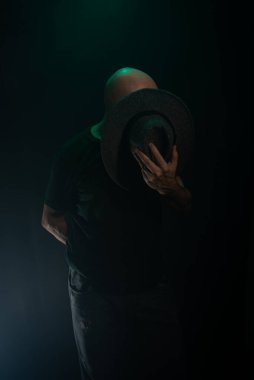 Suratının önünde şapka tutan gizemli bir adam kameraya poz veriyor. Stüdyo portresi. Yapay dumanlı koyu arkaplan.