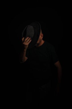 Suratının önünde şapka tutan gizemli bir adam kameraya poz veriyor. Stüdyo portresi. Yapay dumanlı koyu arkaplan.