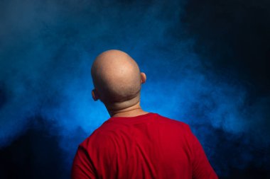 Sakallı, kel, orta yaşlı, sırtı kameraya dönük bir adam. Stüdyoda fotoğraf. Yapay dumanlı mavi stüdyo arka planı.