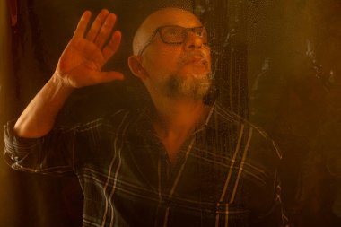 Sarı ışıkla aydınlatılmış gözlüklü sakallı kel bir adam. Şeffaf bir camın içinden fotoğraf için poz veriyor. Koyu arkaplanda izole.