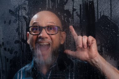 Islak camın arkasında gözlüklü sakallı kel bir adamın komik hareketler yaptığı yakın plan portresi. Siyah arkaplanda izole.