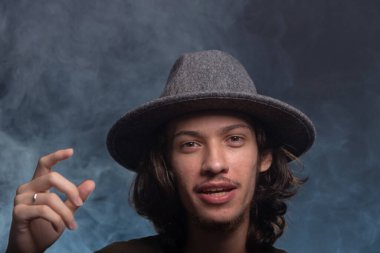 Gri şapkalı ve fotoğraf için poz veren yakışıklı bir gencin yakın plan portresi. Dumanlı siyah arkaplanda izole edilmiş.