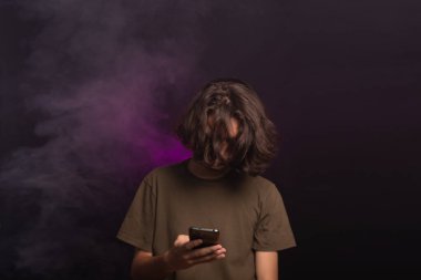 Uzun saçlı, kafası aşağıda cep telefonuna bakan yakışıklı bir gencin portresi. Dumanlı pembe arkaplanda izole edilmiş.