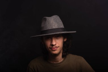 Uzun saçlı, gri şapkalı, fotoğraf için poz veren yakışıklı bir gencin portresi. Siyah arkaplanda izole.