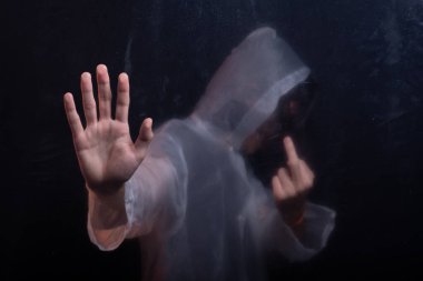 Camların arkasında şeffaf plastik yağmurluk giyen genç bir adam kameraya el hareketi yapıyor. Yüzü gizli. Stüdyo portresi. Koyu arkaplanda izole.