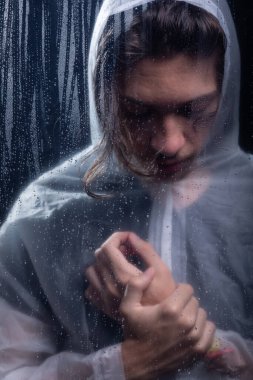 Islak bardağın arkasında elini tutan yağmurluk giyen genç bir adam. Stüdyo portresi. Koyu arkaplanda izole.