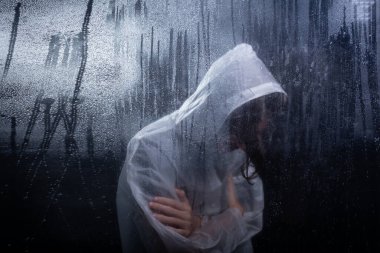 Yağmurluk giyen genç adam ıslak camın arkasında kollarını kavuşturmuş üşüyormuş gibi hissediyordu. Stüdyo portresi. Koyu arkaplanda izole.