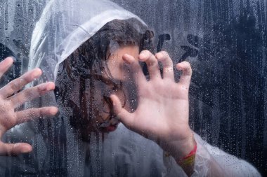 Islak camın arkasında eli görünen yağmurluk giyen bir adamın yakın plan portresi. Stüdyo portresi. Koyu arkaplanda izole.