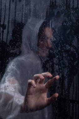 Islak camın arkasında yağmurluk giyen insan portresi. Elinle cama dokunuyorsun. Stüdyo portresi. Koyu arkaplanda izole.