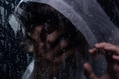 Islak camın arkasında eli yüzünde yağmurluk giyen bir adamın yakın plan portresi. Stüdyo portresi. Koyu arkaplanda izole.