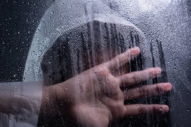 Islak camın arkasında yağmurluk giyen ve elini yüzünü kapatan bir adamın yakın plan portresi. Stüdyo portresi. Koyu arkaplanda izole.