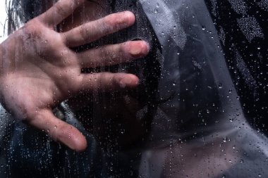 Islak camın arkasında yağmurluk giyen ve elini yüzünü kapatan bir adamın yakın plan portresi. Stüdyo portresi. Koyu arkaplanda izole.