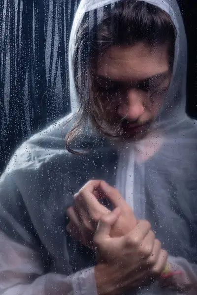 Islak bardağın arkasında elini tutan yağmurluk giyen genç bir adam. Stüdyo portresi. Koyu arkaplanda izole.