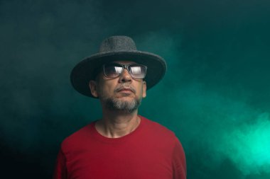 Sakallı adam şapka takıyor ve fotoğraf için poz veriyor. Stüdyoda portre. Yapay dumanlı yeşil arkaplan.