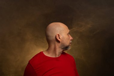 Kırmızı gömlek giyen kel sakallı adam yana bakıyor. Stüdyo portresi. Yapay dumanlı sarı arkaplan.
